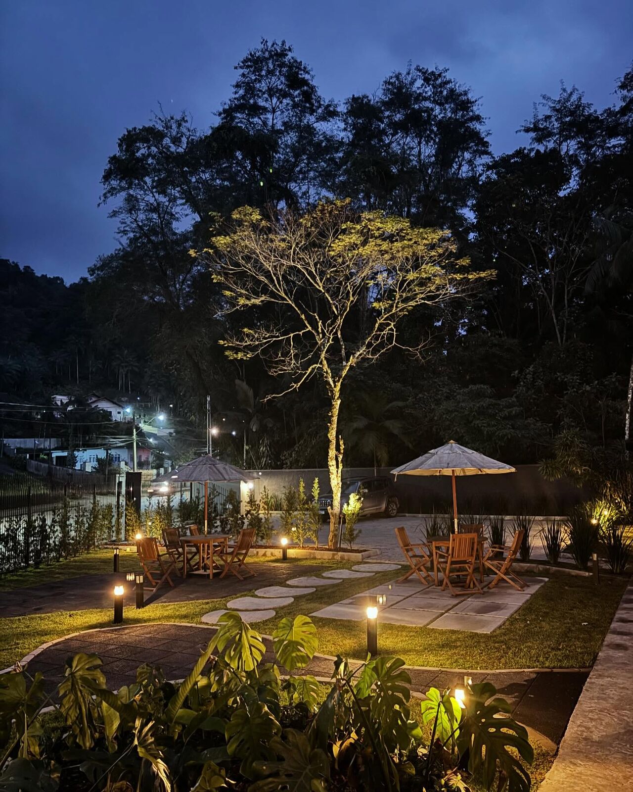 Jardim da Pommerlofts ao entardecer, com mesas, guarda-sóis e caminhos iluminados entre as árvores